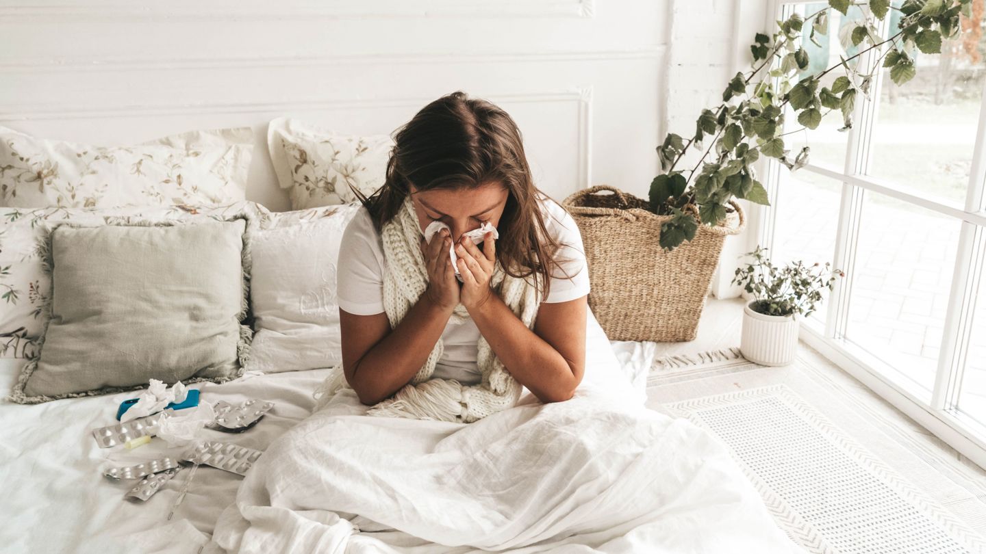 Bildet viser en kvinne som sitter i senga og snyter seg.